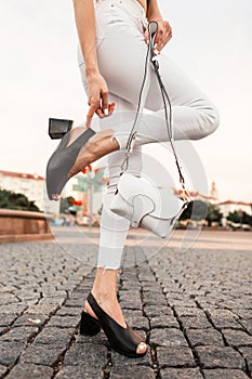 Female legs in white jeans in black leather sandals with a bag on a stone road in the city. Close-up