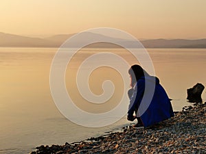 Female looking sunset on sea, lake. Woman sitting on rocks. Lonely person. Girl alone outside. Vacation on sea. Relax