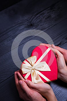 Female and male caucasian hands holding gift box in shape of heart on black wooden background