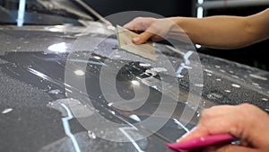 A female and a male hand with plastic scrapers glue protective film on new car.