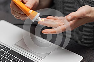 Female or male hands applying hand cream on fingers to moisturize skin while working at desk in home office near laptop computer