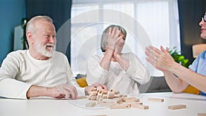 female and male pensioners have fun playing board game with nurse sitting at table in room