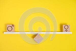 Female and male symbols are depicted on wooden cubes balanced scales yellow background.