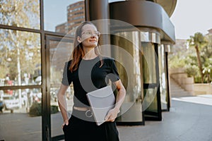 Female manager in eyeglasses holding laptop while standing on modern building background