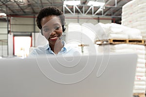 Female manager talking on headset while using laptop at desk in warehouse