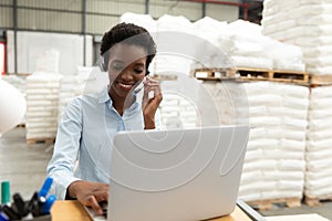 Female manager talking on headset while using laptop at desk in warehouse