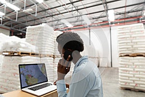 Female manager talking on mobile phone while using laptop at desk in warehouse