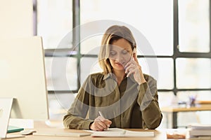 Female manager is talking on phone and taking notes in notebook while sitting at her desk in office.