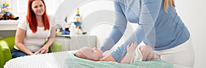 Female massage therapist or a doctor examining newborn baby boy with the mother watching in the background. Baby massage banner.