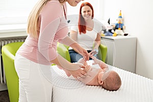 Female massage therapist or a doctor examining newborn baby boy with the mother watching in the background. Baby massage concept.