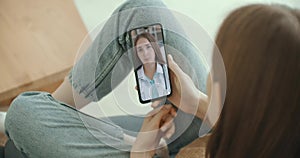 Female medical assistant wears white coat, headset video calling distant patient on smartphone. Doctor talking to client