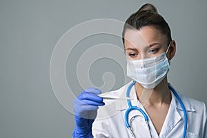 A female medical worker in a white robe and mask holds and looks at an electronic thermometer.