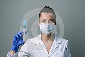 A female medical worker in a white robe and mask holds a syringe.