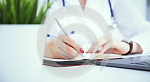 Female medicine doctor hand holding silver pen writing something on clipboard and working laptop closeup. Physician