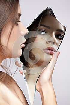 Female with mirror shard in hand posing on gray background. Face reflection in mirror splinter