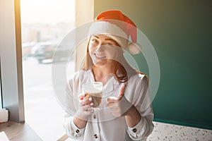 Female model in christmas hat shows thumb up. Young woman drink coffee and smile.