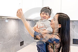 Female mother of east asian ethnicity with toddler son attempting to test cabinet door handles, signifying mom life and stay-at-