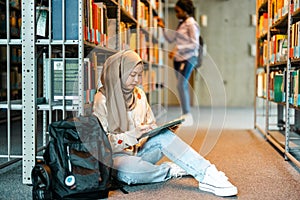 Female muslim student sitting on floor in library and reading book