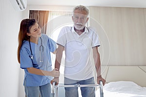 Female nurse or doctor helping elderly patient man lean to walk with orthopedic walker, patient practice walking inside house with