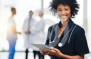 Female nurse or doctor holding a tablet with colleagues standing in the background. Confident, smiling healthcare