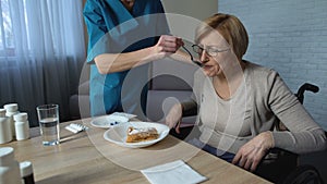 Female nurse helping disabled old lady to eat, medical support and care, sadness