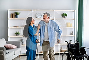 Female nurse helping elderly male to walk with crutches at home. Professional medical care for disabled older patients photo