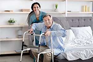 Female nurse helping elderly male with walking frame stand up from bed at home. Professional care for disabled patients