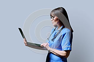 Female nurse using laptop, profile view on gray studio background