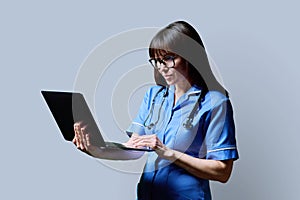 Female nurse using laptop, profile view on gray studio background