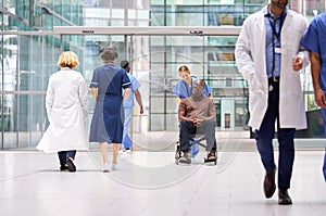 Female Nurse Wearing Scrubs Wheeling Patient In Wheelchair Through Lobby Of Modern Hospital Building