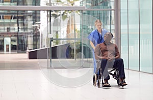 Female Nurse Wearing Scrubs Wheeling Patient In Wheelchair Through Lobby Of Modern Hospital Building