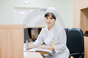 A female nurse in a white medical gown sits at a wooden desk at the reception desk and makes entries in a work log. Woman doctor