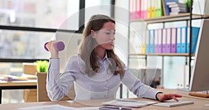 Female office worker doing exercises with dumbbells in a modern office in front of laptop computer