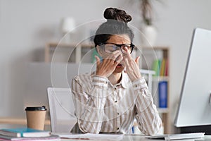 Female office worker in glasses rubbing tired eyes, exhausted from overworking, sitting at workplace in office