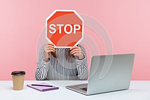 Female office worker in striped shirt sitting at workplace hiding face behind red stop traffic sign avoiding conflicts, afraid of