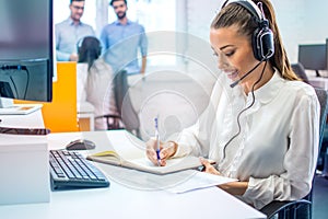 Female operator with headset writing notes while talking to client in customer support center