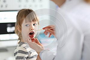 Female paediatrician examining little kid patient throat with wooden stick