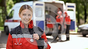 Female paramedic smiling into camera, ambulance crew blurred on background