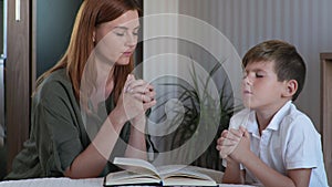 Female parent with male child kneel by bed with folded hands and pray to God