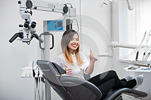 Female patient with perfect white teeth sitting in dental chair, smiling and showing thumbs up after treatment