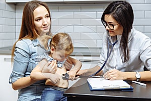 Female Pediatrician examining little girl with stethoscope. Mother holding her kid. Doctor visit his patient at home