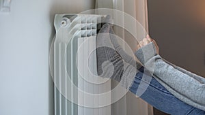 Female person putting feet in grey socks on heating radiator tries to warm up in cold apartment