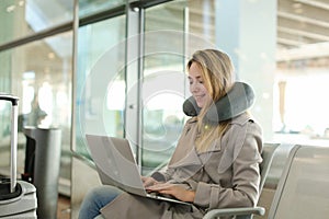 Female person sitting in waiting room with laptop near valise, using neck pillow.