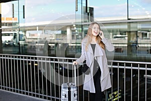 Female person standing with valise and talking by smartpone near airport.