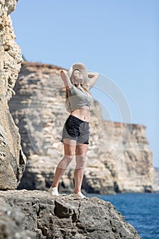 Female person in wide-brimmed hat stands with hands behind head among rocks