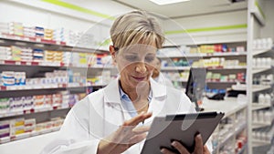 Female pharmacist adjusting settings on digital tablet while assistant sorts out scripts behind counter in pharmacy