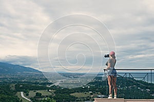 A female photographer in a red cap with a camera stands on the balcony opposite of the Greek city of Katerini at sunset. Katerini