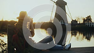 Female photographer sits on a sunset lake pier. Girl with camera takes a photo of traditional old rustic windmill. 4K.