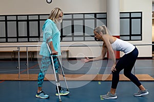 Female physiotherapist assisting disabled senior woman walk with elbow crutches in sports center