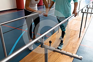 Female physiotherapist helping disabled senior woman walk with parallel bars in sports center